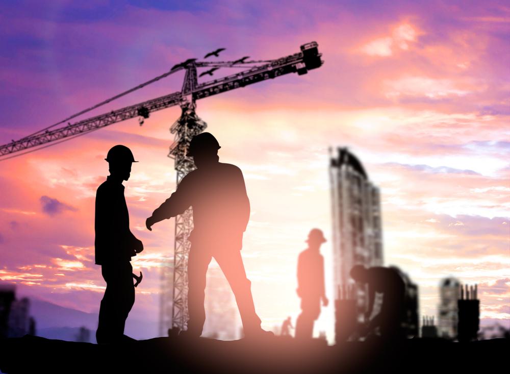 Photo of a silhouette of construction workers and a tower crane against a vibrant purple and orange sunset sky.